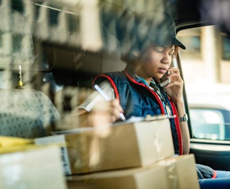 black-female-courier-communicating-mobile-phone-taking-notes-while-sitting-delivery-van-view-is-through-window_637285-2208